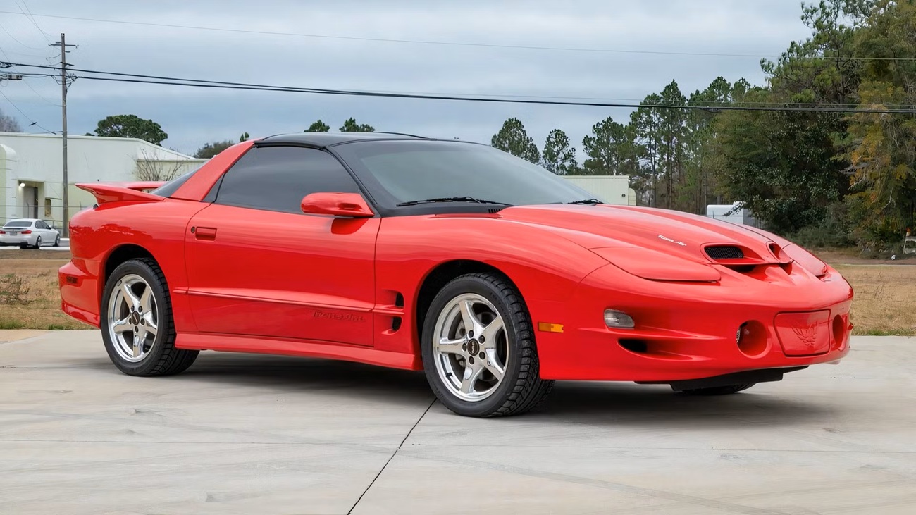 Red Pontiac Firebird Trans Am sports coupe with a spoiler, parked on a concrete driveway with tinted windows.