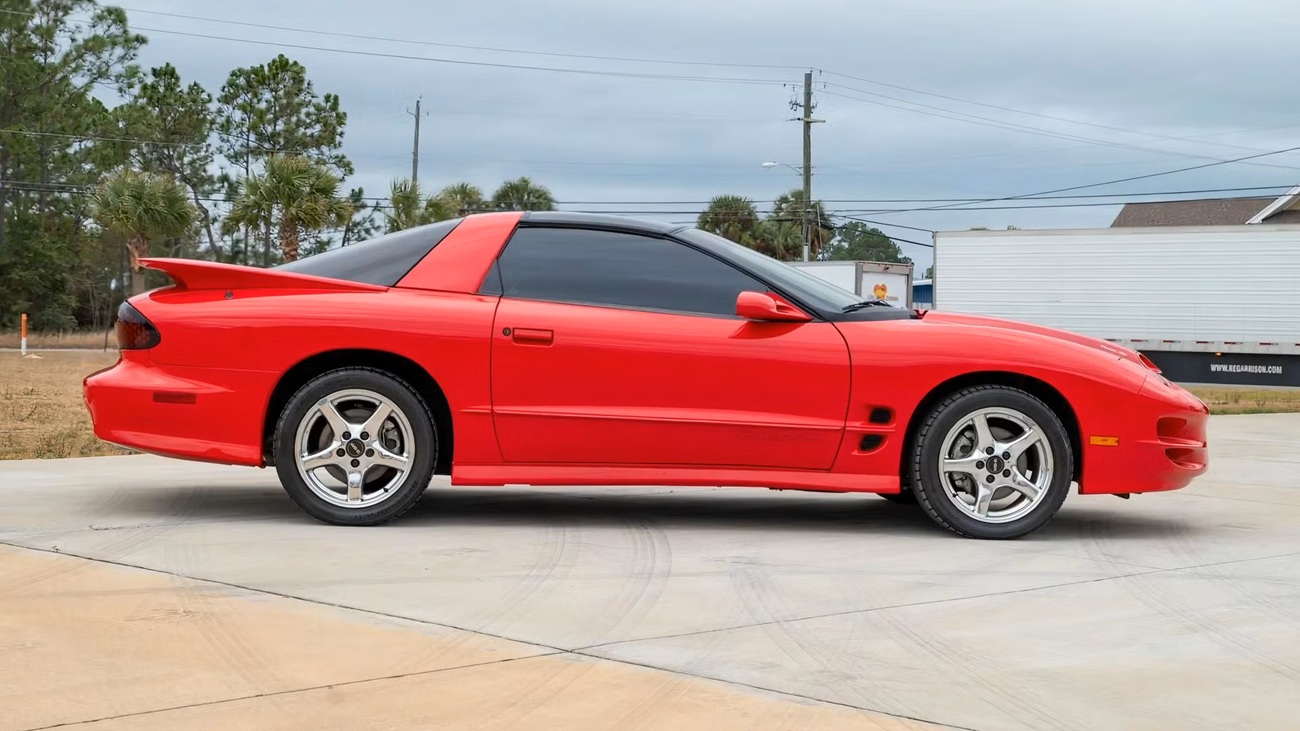 Side view of a bright red sports car parked on a light concrete driveway, tinted windows and a spoiler visible at the rear.