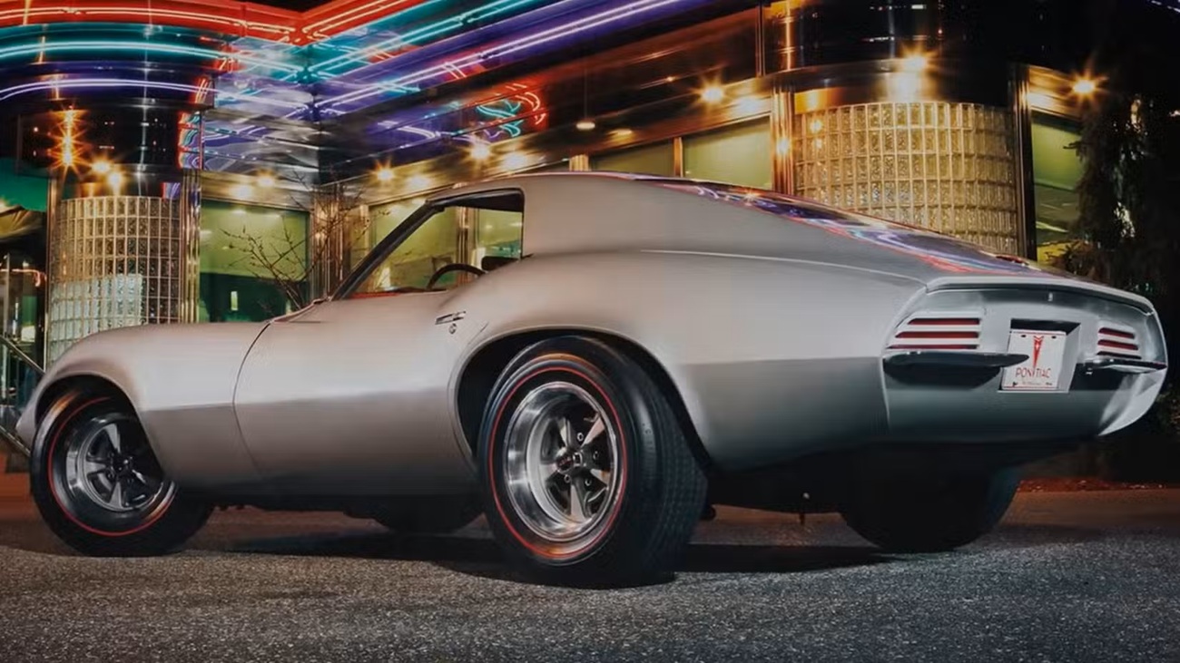 Rear three-quarter view of a silver classic Pontiac sports car with chrome wheels at night, under neon lights.