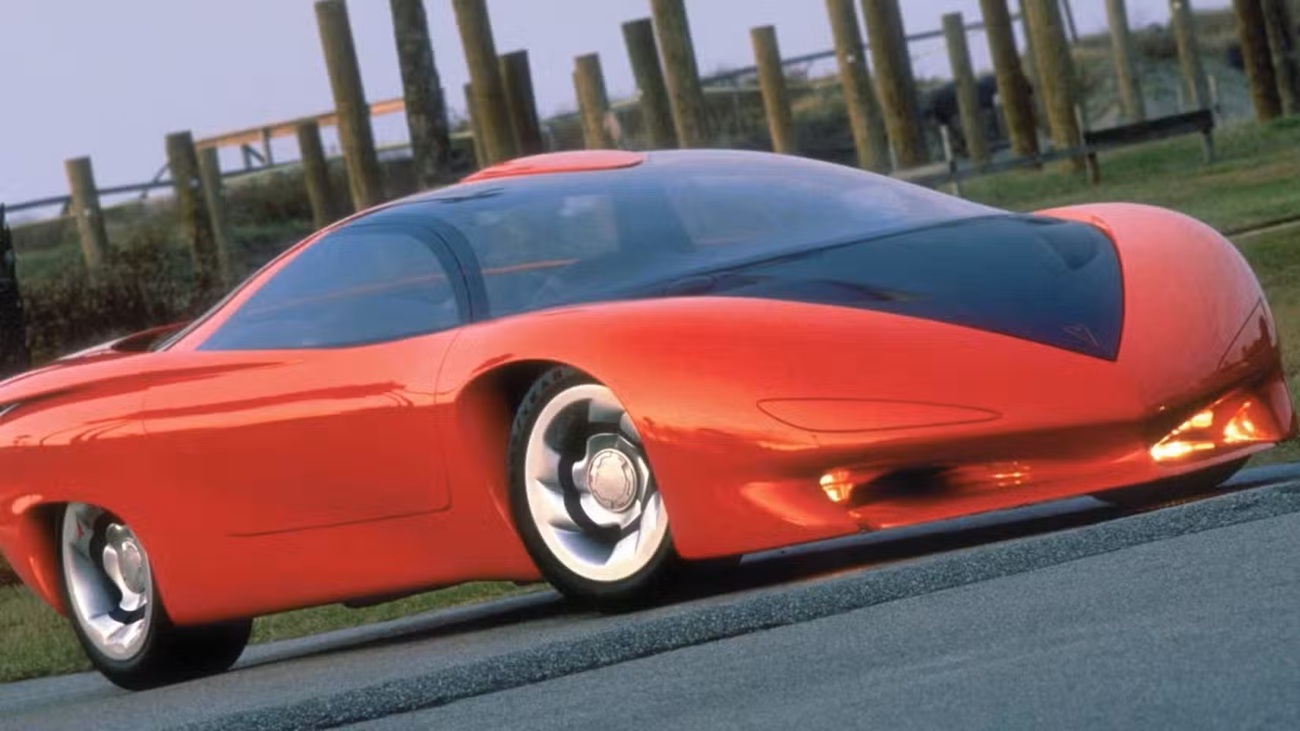 Red futuristic sports car with a black tinted canopy-style windshield on a road, urban background behind a fence.
