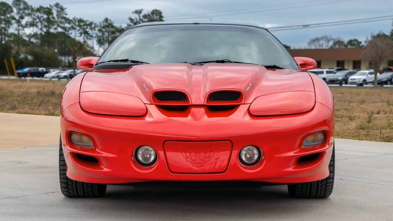 Front view of a red Pontiac sports car with dual hood scoops and round headlights, parked on a concrete surface with a grassy area in the background.