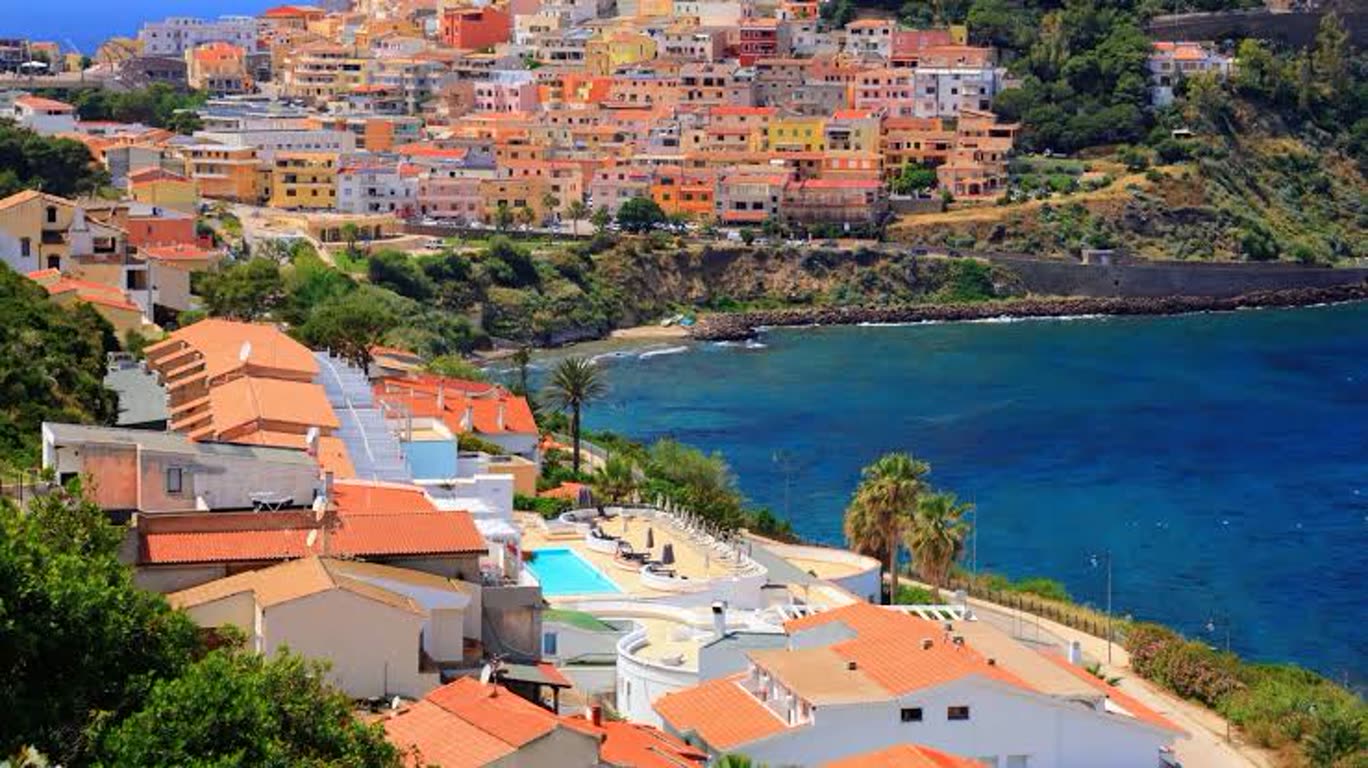 Colorful coastal village on a hillside overlooking a blue bay with orange-tiled roofs and a foreground pool area.
