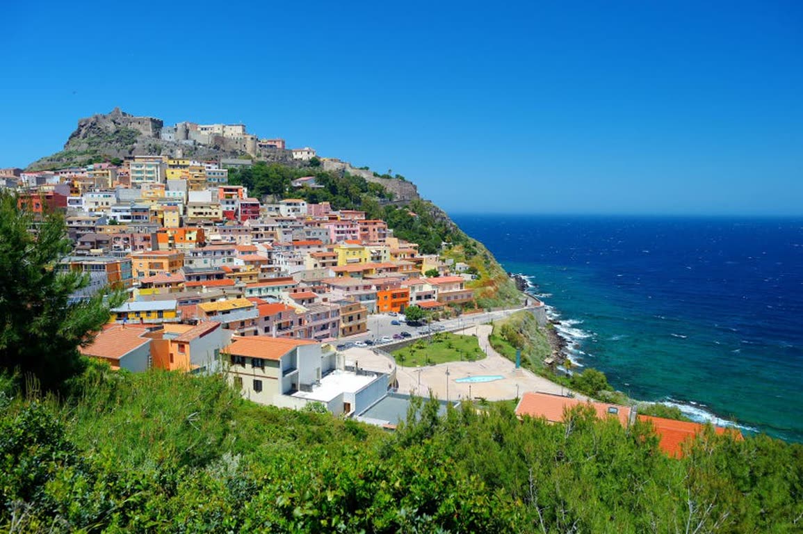 Colorful hillside town perched on a cliff above a blue sea under a clear sky, with a fortress at the top edge.