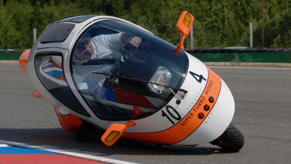 Orange-and-white three-wheeled race car leaning into a corner on a track, cockpit with a driver visible through the canopy.