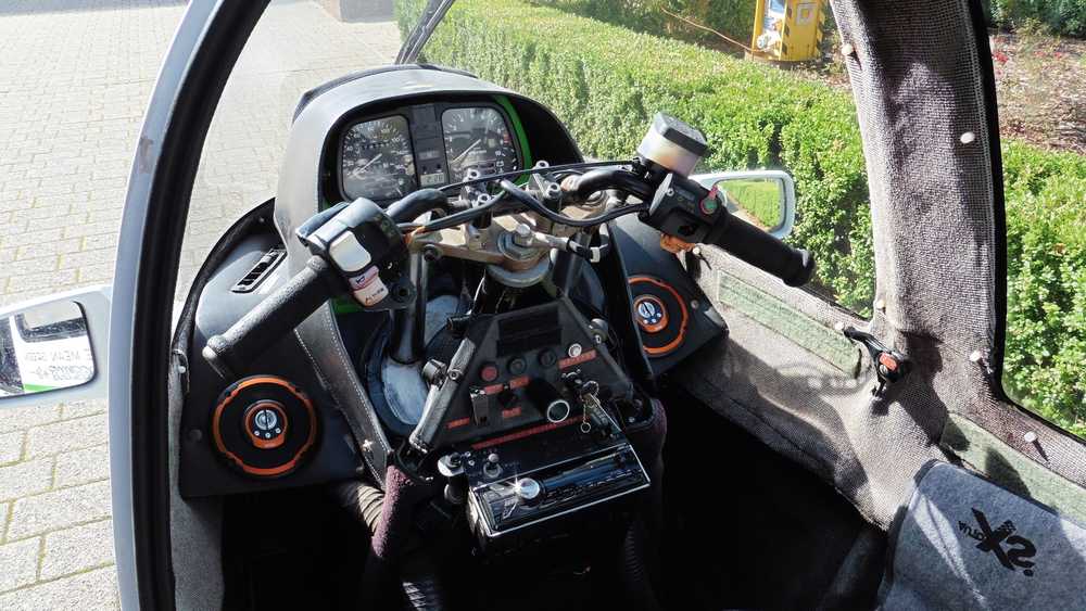Cockpit of a small aircraft with dual control sticks and instrument panel, seen from the pilot's seat, with a paved area outside and greenery nearby.