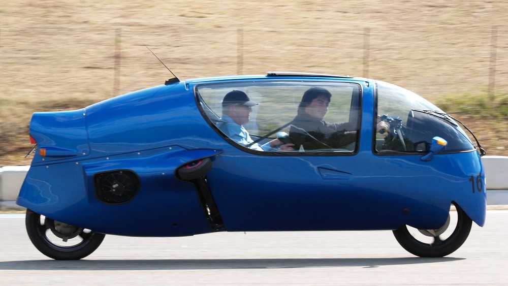 Blue three-wheeled car with two people inside driving on a road, in motion.