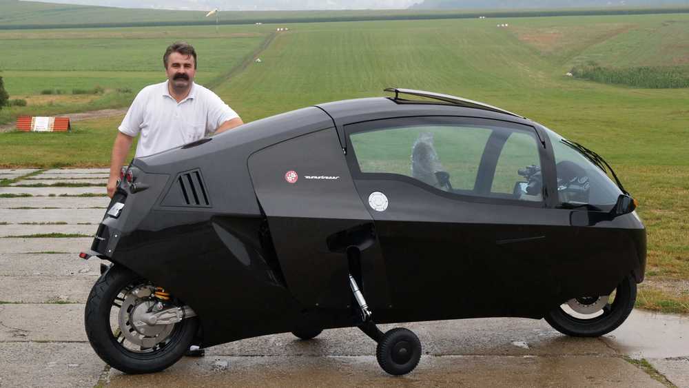 Man standing beside a sleek black enclosed three-wheeled motorcycle on an airstrip, with green fields in the background.