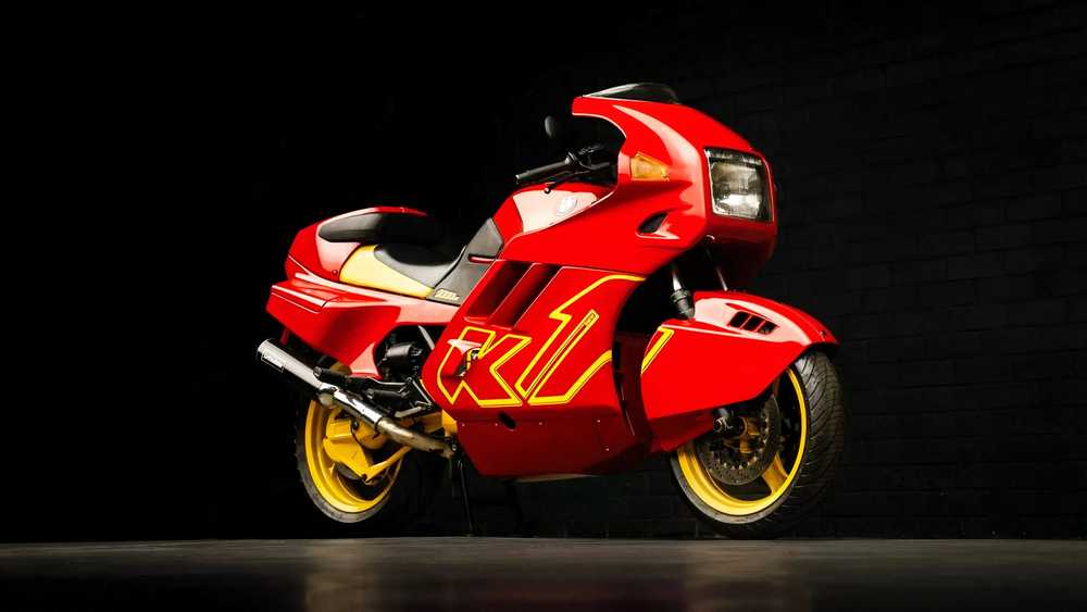 Red sport motorcycle with yellow rims shown from a side angle against a dark studio backdrop.