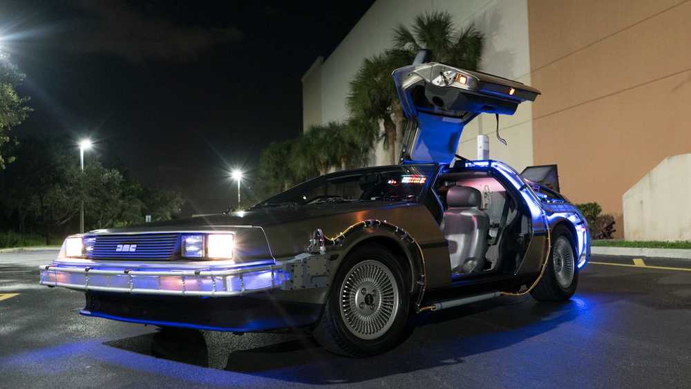DeLorean DMC-12 time machine with gull-wing doors open, blue underglow, parked in a dim night parking lot.
