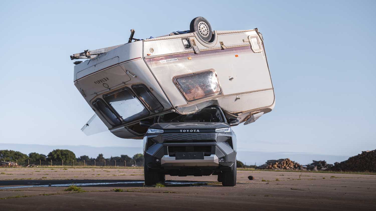 Toyota pickup truck balances an oversized camper on its front end, parked on an open, paved area with a clear blue sky behind.