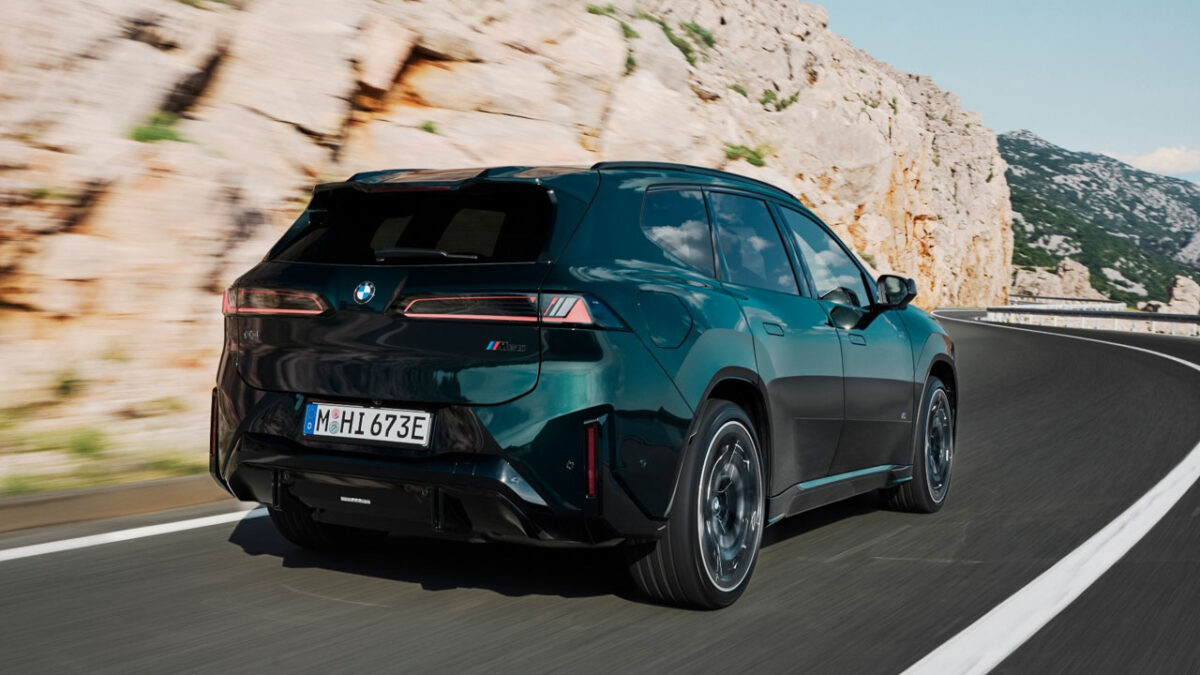 نمای سه‌رخ عقب خودروی برقی BMW iX3 M60 xDrive سبز تیره با رینگ‌های اسپرت، چراغ‌های کشیده عقب و دیفیوزر مشکی در جاده‌ای کوهستانی