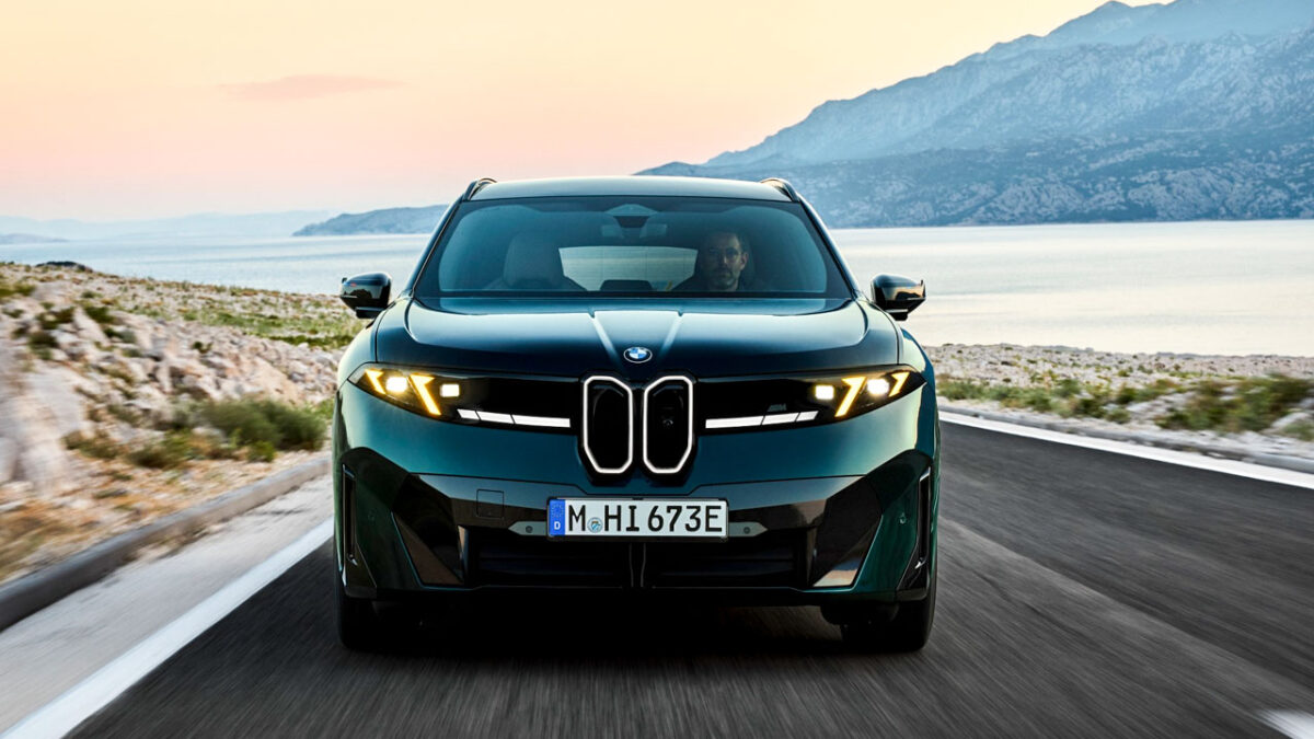 نمای روبه‌روی BMW iX3 M60 xDrive سبز تیره با چراغ‌های زرد اختصاصی M، جلوپنجره نورانی و سپر اسپرت در جاده‌ای ساحلی هنگام غروب