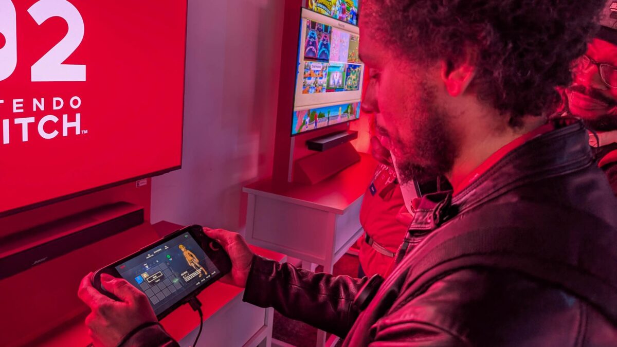 شرکت‌کننده‌ای در یک رویداد عمومی با نورپردازی قرمز، در حال امتحان کردن Nintendo Switch 2 در حالت دستی است که صفحه‌نمایشی رنگی از منوی بازی را نشان می‌دهد و بنر بزرگ برند در پس‌زمینه دیده می‌شود.