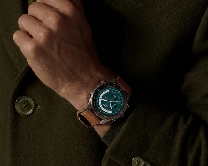 نمای نزدیک از مچ دست فردی با لباس سبز، که ساعت هوشمند Garmin با بند چرمی قهوه‌ای و صفحه نمایش عقربه‌ای دیجیتال را به دست بسته است.