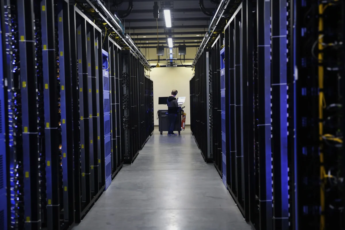 نمای سراسری از راهروی یک مرکز داده یا سرور روم (Data Center) بزرگ با رک‌های سرور سیاه‌رنگ و نورپردازی آبی، در حالی که یک تکنسین در حال کار است.