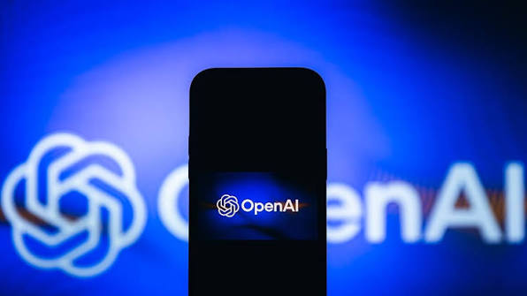 گوشی هوشمند در نمای روبه‌رو با صفحه سیاه که لوگوی درخشان OpenAI روی نمایشگر آن باز شده و در پس‌زمینه‌ای با نور آبی عریض قرار دارد.
