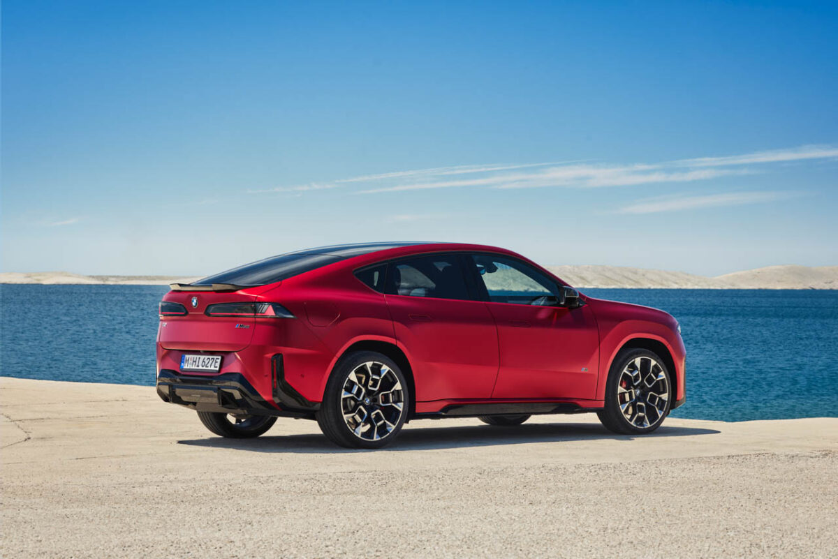 نمای سه‌رخ عقب خودروی برقی BMW iX4 M60 قرمزرنگ در کنار دریا، با سقف شیب‌دار، چراغ‌های کشیده عقب و رینگ‌های اسپرت