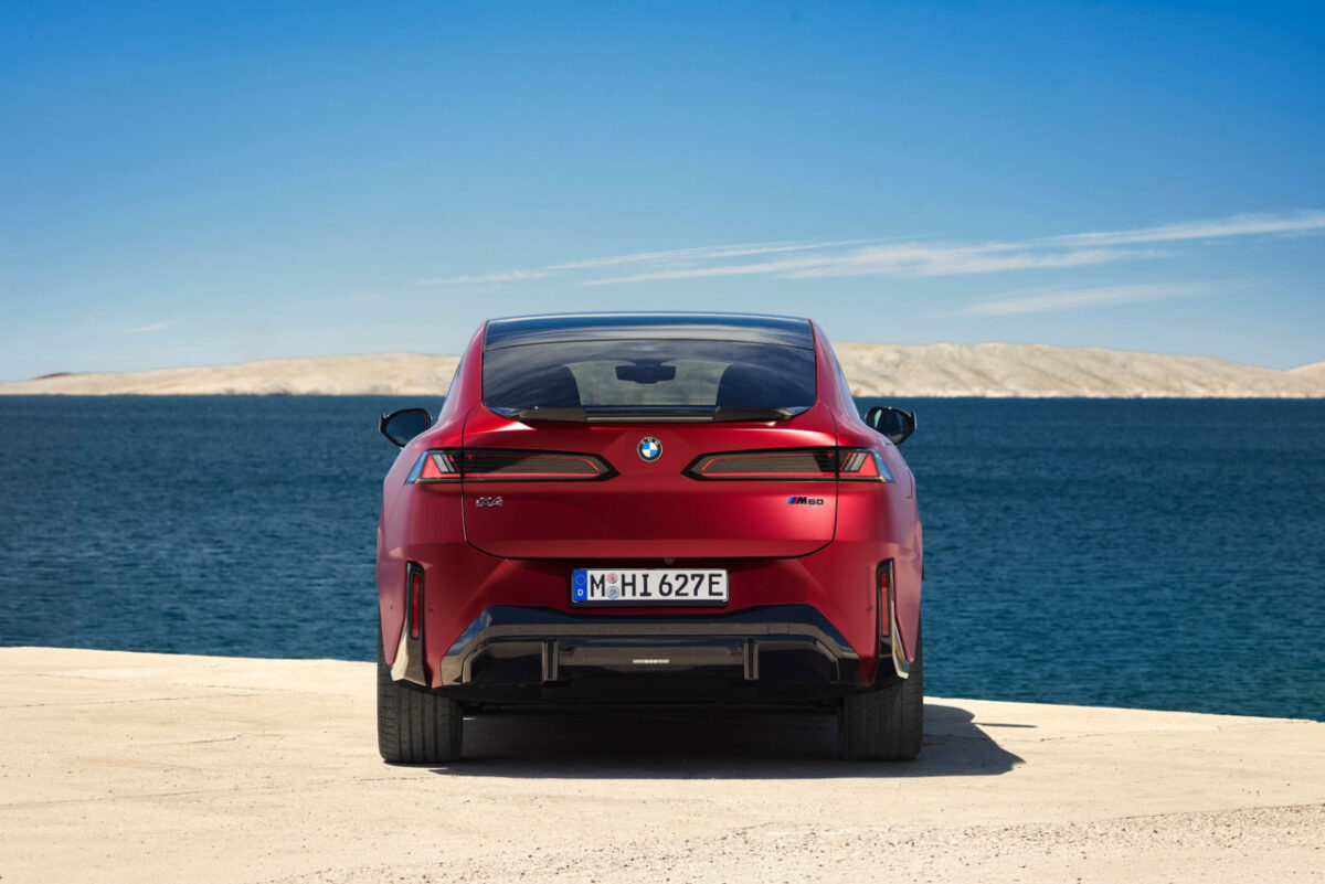 نمای مستقیم عقب شاسی‌بلند برقی BMW iX4 M60 با چراغ‌های افقی باریک، نشان BMW و سپر اسپرت در کنار دریا