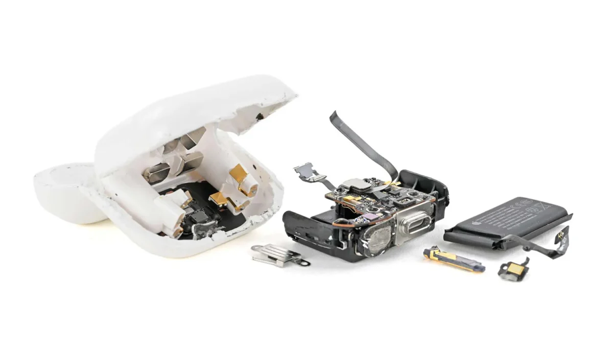 عکس جداسازی قطعات (teardown) جعبه شارژ AirPods 5 توسط iFixit روی پس‌زمینه سفید. پوسته سفید جعبه شکافته شده و قطعات داخلی شامل مگنت‌ها و پین‌های شارژ، برد الکترونیکی با درگاه USB-C و کابل فلکسی بلند، باتری مشکی مستطیلی و چند قطعه فلزی کوچک کنار هم چیده شده‌اند.