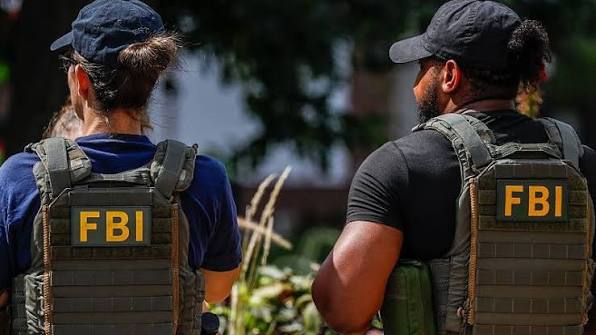 دو مأمور FBI از پشت، با جلیقه‌های تاکتیکی دارای نشان زرد FBI، در فضای باز و کنار پوشش گیاهی.