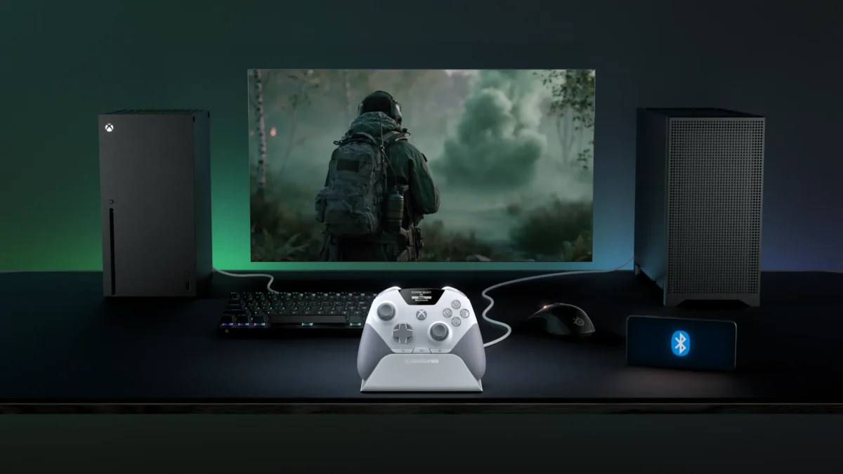 میز گیمینگ تیره با کنترلر سفید خاکستری SteelSeries Aeon Pro روی پایه شارژ اختصاصی در مرکز؛ کنسول مشکی Xbox Series X با نور سبز در سمت چپ، کیس کامپیوتر مینی در سمت راست، مانیتور در حال نمایش بازی جنگی شوتر اول‌شخص، کیبورد مکانیکی، ماوس گیمینگ و گوشی نمایش‌دهنده لوگوی بلوتوث.