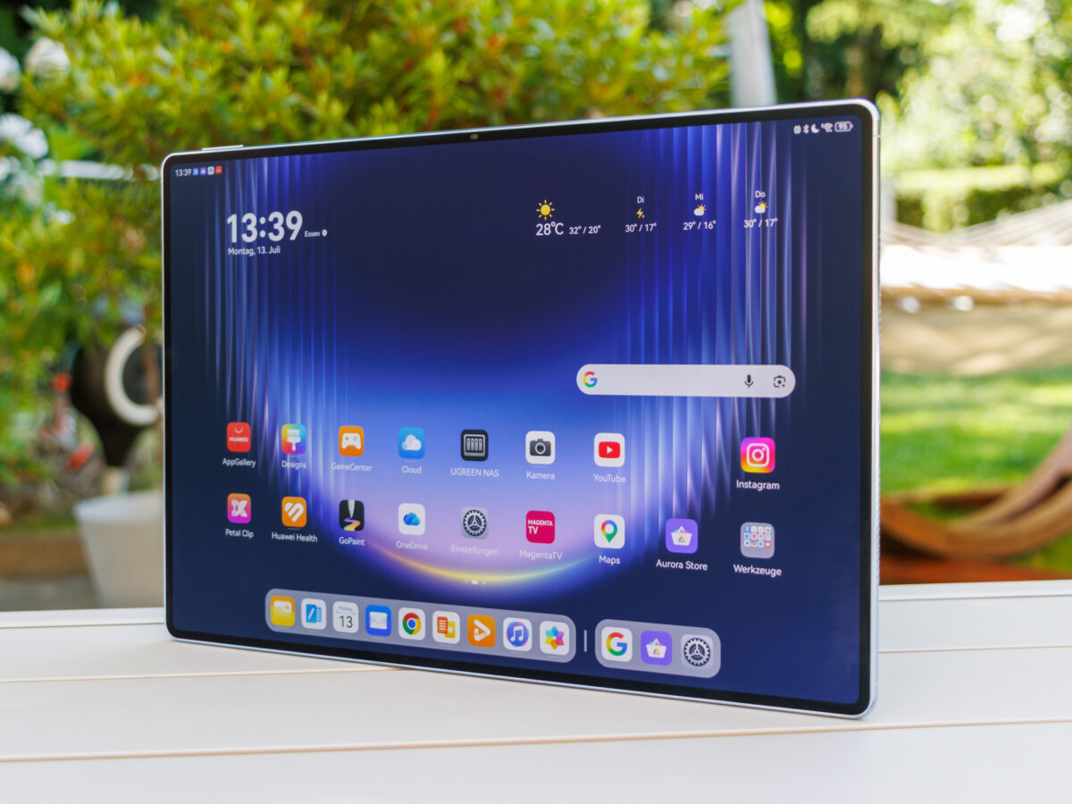 نمای روبه‌رو تبلت هواوی MatePad Pro Max با نمایشگر بزرگ، رابط کاربری HarmonyOS، صفحه اصلی شامل ساعت ۱۳:۳۹، ابزارک هوا و آیکون‌های اپلیکیشن‌هایی مانند یوتیوب و اینستاگرام، روی میز چوبی سفید در فضای باز.