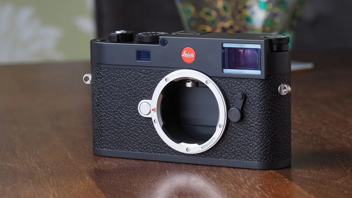 دوربین عکاسی دیجیتال لایکا (Leica) با بدنه‌ی مشکی چرمی و مانت لنز فلزی بدون لنز، قرارگرفته روی سطح چوبی.