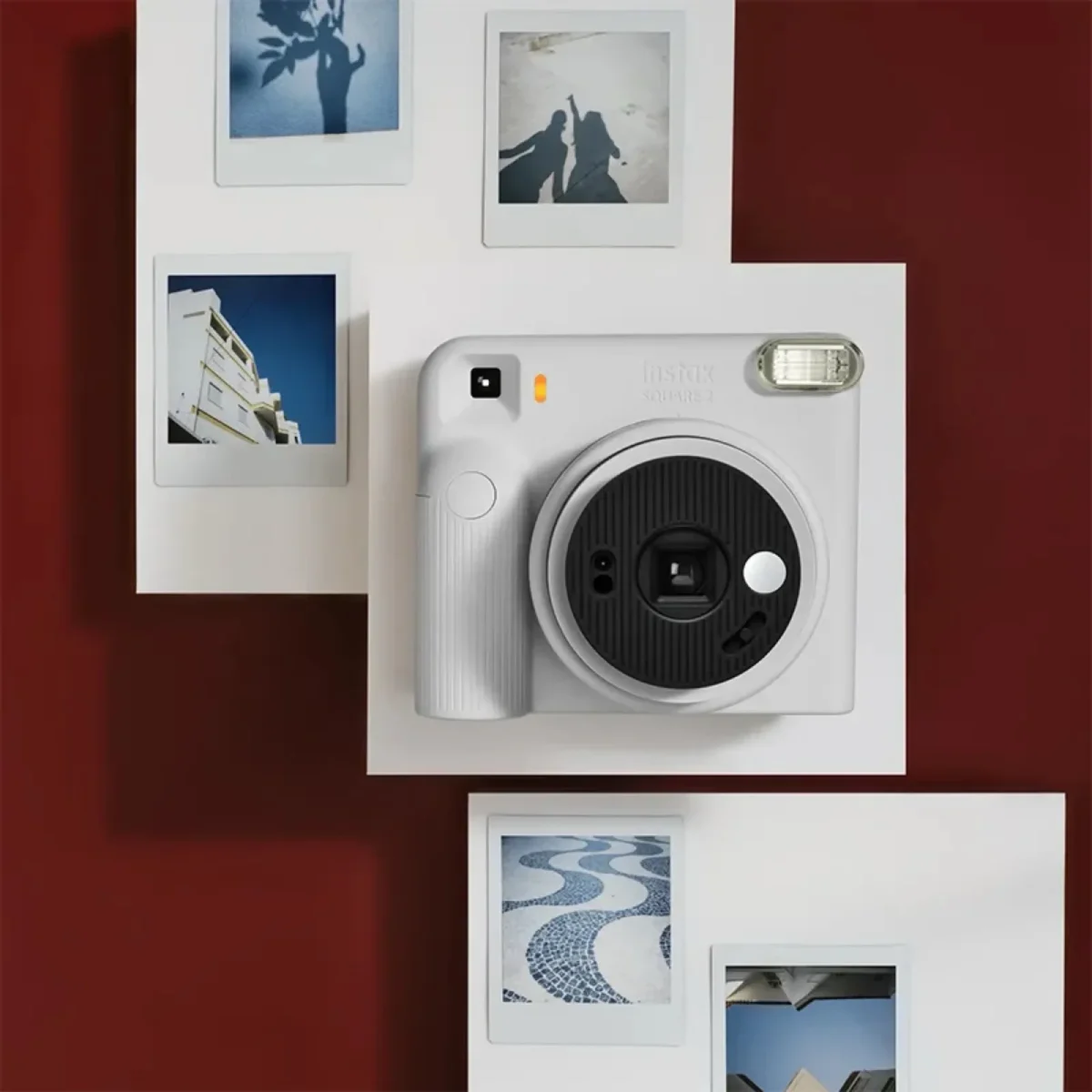 دوربین چاپ سریع سفید رنگ Instax Square SQ2 با لنز مشکی و فلاش نقره‌ای در مرکز تصویر، احاطه‌شده توسط چهار عکس چاپ‌شده مربعی روی پس‌زمینه زرشکی.