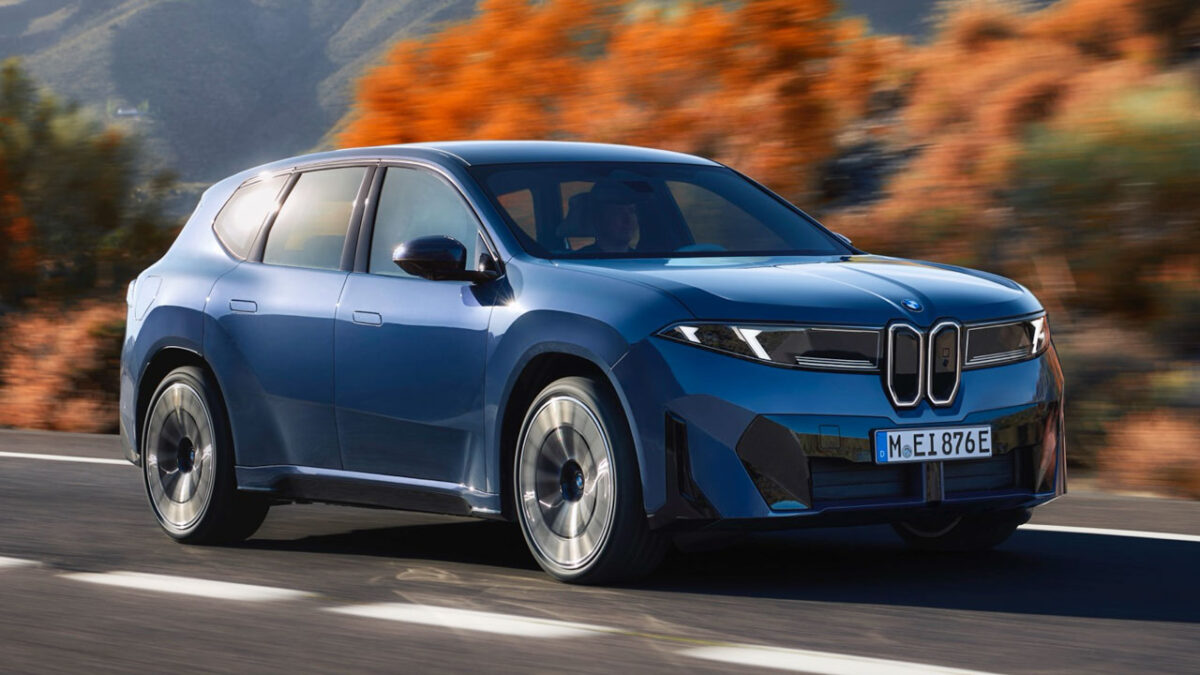 BMW iX3 50 xDrive: شاسی‌بلند برقی آبی BMW iX3 50 xDrive در حال حرکت در جاده با پس‌زمینه پاییزی