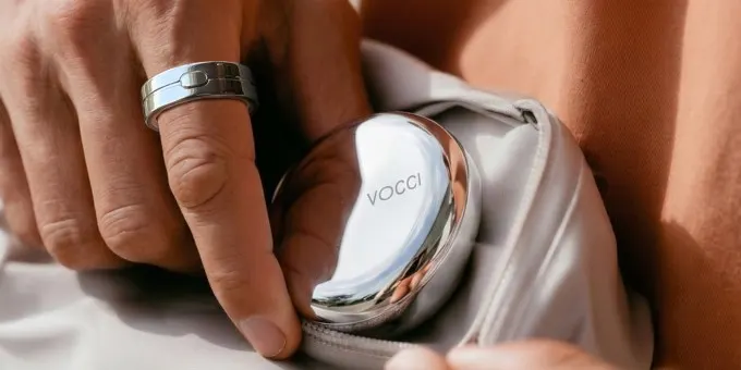 دست مردی که انگشتر نقره‌ای برند Vocci را در انگشت کرده و یک محفظه فلزی براق دایره‌ای شکل با برچسب Vocci را در کیف پولی سفید جای داده؛ محصولی ترکیبی از جواهر و کیف پول هوشمند با نور طبیعی گرم.