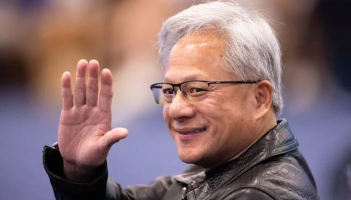 جن‌سن هوانگ (Jensen Huang)، مدیرعامل شرکت انویدیا، با ظاهر شناخته‌شده‌اش شامل موهای نقره‌ای و عینک، در حالی که کت چرمی مشکی به تن دارد و در یک رویداد رسمی برای مخاطبان دست تکان می‌دهد.