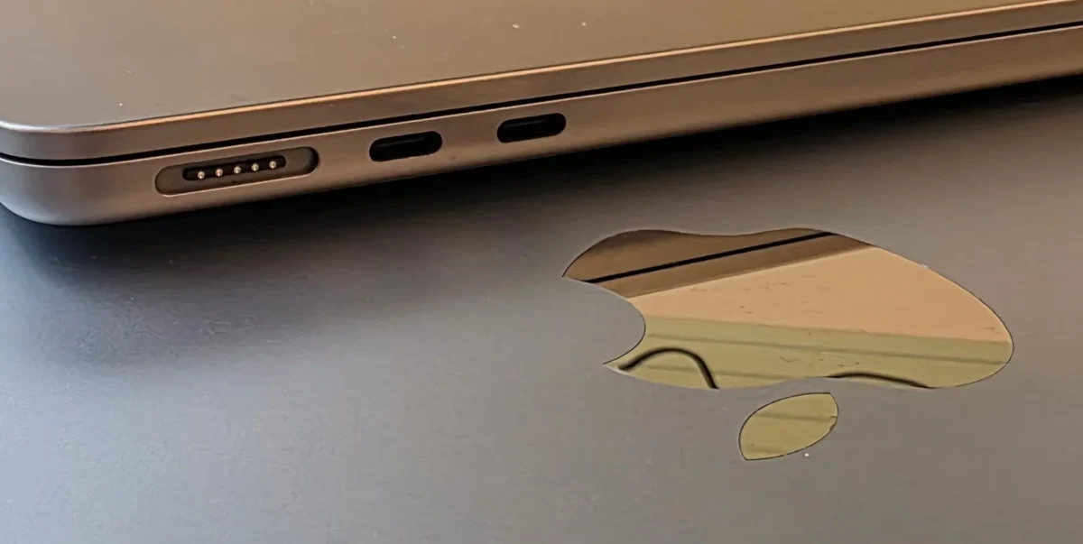 نمای کلوزآپ از لبه کناری یک لپتاپ اپل به رنگ طلایی-قهوه‌ای که پورت MagSafe و دو پورت USB-C روی آن دیده می‌شود، در کنار سطح مشکی با لوگوی برجسته اپل که انعکاس محیط در آن پیداست.