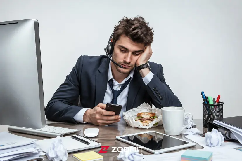 مردی خسته با هدست که در حال چک کردن گوشی هوشمند در میز کار است