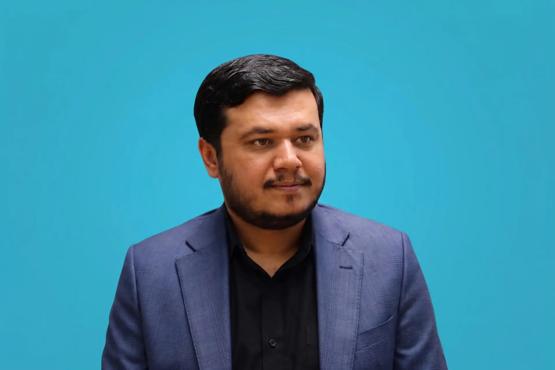 مرد جوان با کت سرمه‌ای و پیراهن مشکی در مقابل پس‌زمینه آبی