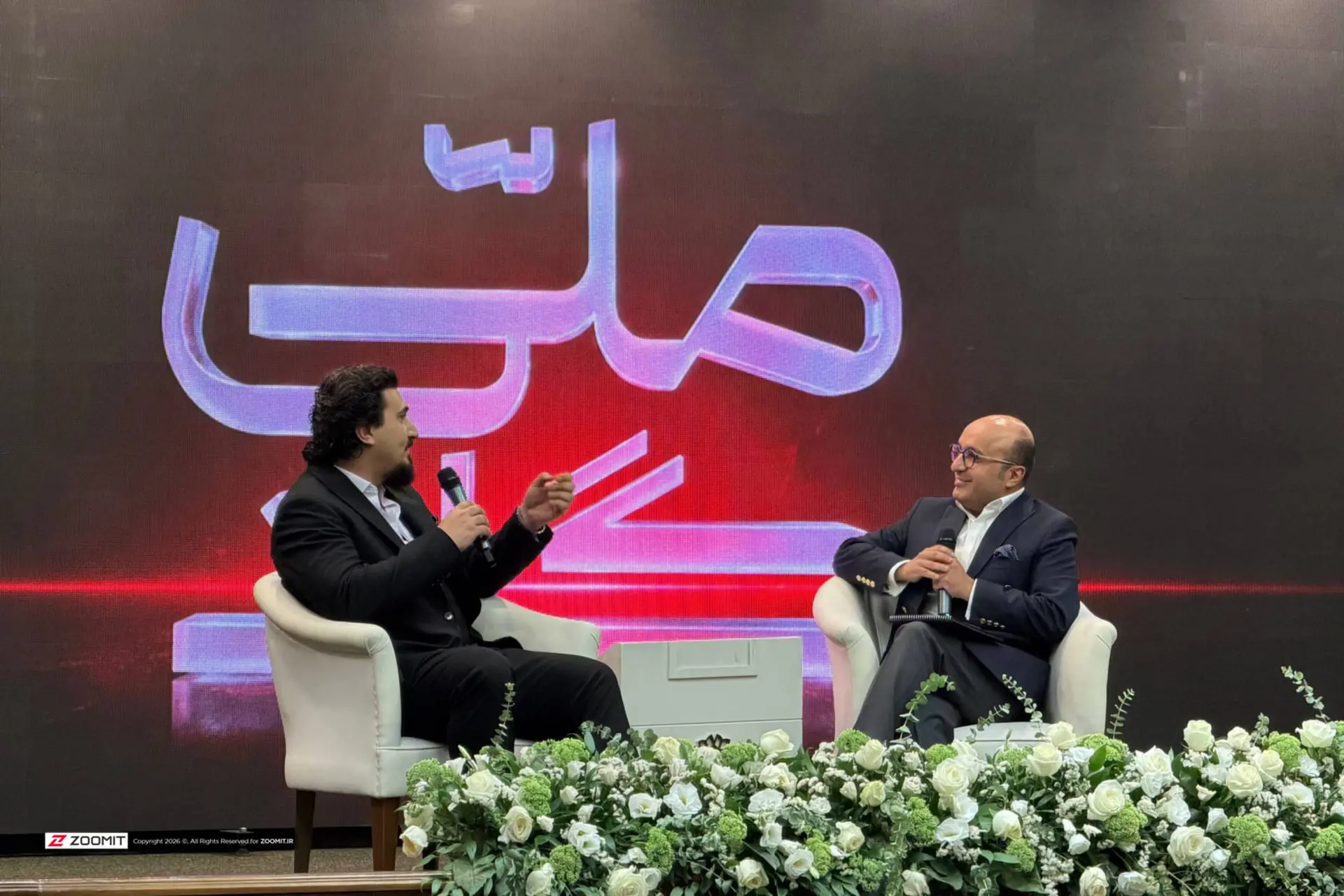 دو مرد در حال گفت‌و‌گو در یک نشست رسمی با پس‌زمینه لوگوی بزرگ