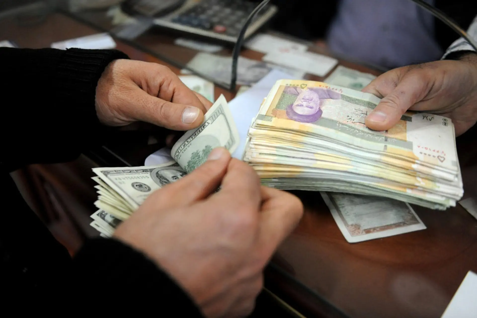 دست‌های افراد در حال شمارش اسکناس‌های دلار (US Dollar) و ریال ایران