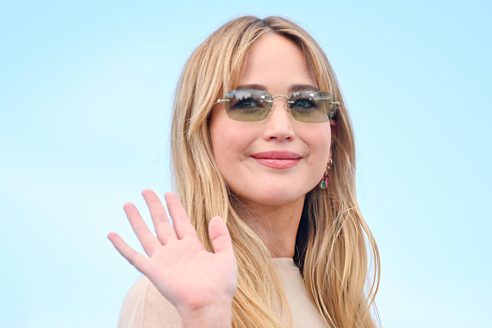 جنیفر لارنس (Jennifer Lawrence) با عینک آفتابی و لبخند در مقابل پس‌زمینه آبی